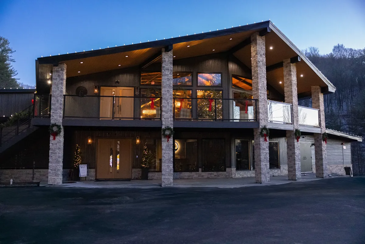 A warmly lit exterior shot of a stone-pillared deck adorned with decorative lights and holiday decorations. Large windows glow invitingly, and the setup suggests a perfect space for gatherings that transition seamlessly from indoor to outdoor. The mountain backdrop is visible, emphasizing the scenic setting.