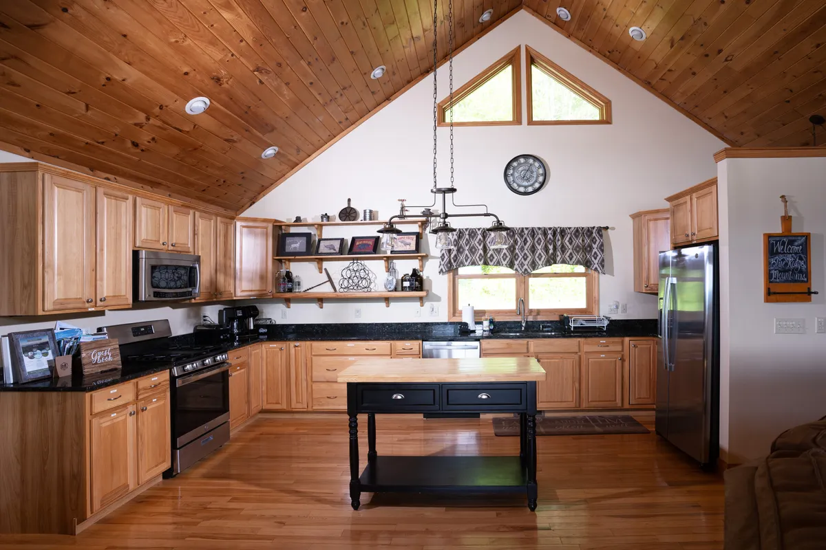A cozy kitchen space with a central island, warm wooden cabinetry, a sleek black countertop, and large windows that flood the room with natural light. The setting suggests a welcoming environment perfect for cooking, gathering, and sharing meals. The expansive windows may offer glimpses of the mountain scenery outside.
