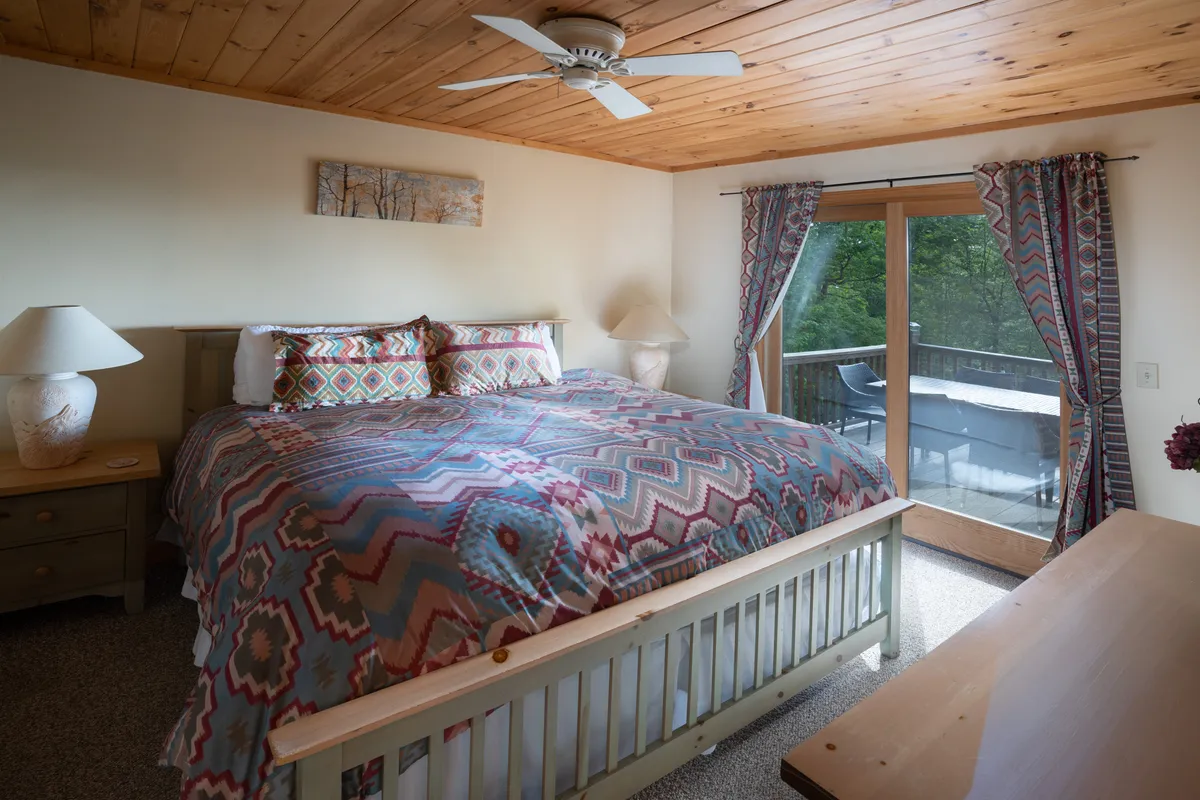 This image shows a cozy bedroom with colorful bedding, a large sliding glass door that opens to natural wooded views, and decorative bedside lamps. The scene is bright and inviting, with the natural light and greenery outside contributing to a sense of peaceful seclusion.
