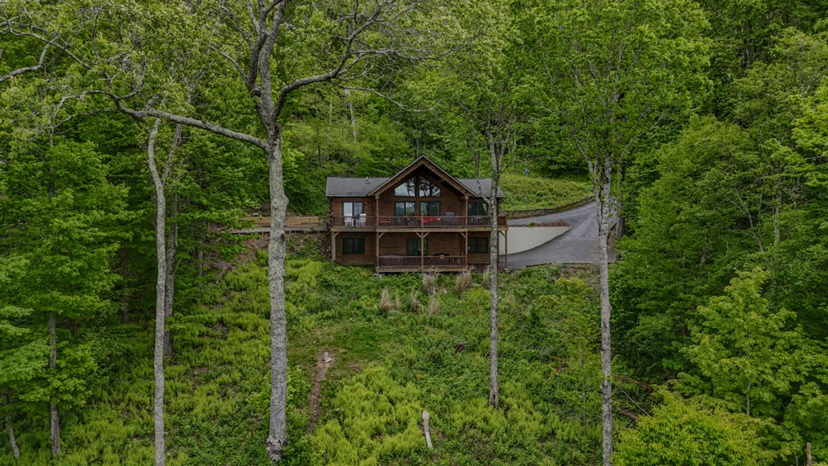 A picturesque wooden cabin sits nestled among tall, leafy trees, with a driveway curving gently up to the entrance. The calm, late-day light highlights the rustic wood exterior, while the covered porch offers a welcoming space to relax and soak in the natural surroundings.