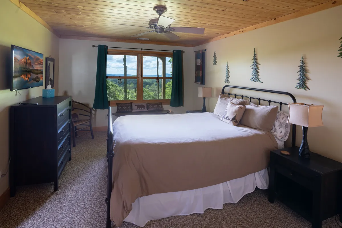 A tranquil bedroom scene with a comfortable bed, a window looking out to the woods, a dresser, and artistic wall décor featuring decorative trees. The soft light and decor create a peaceful, cozy atmosphere ideal for unwinding.
