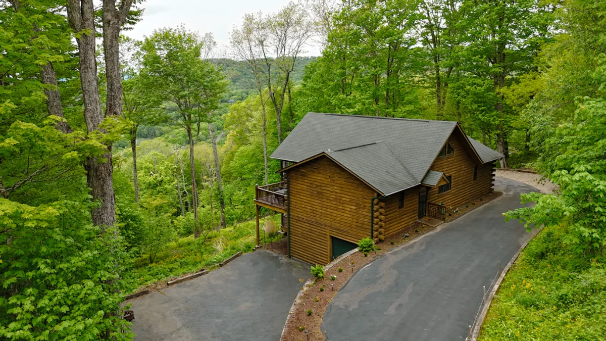 A charming wooden cabin nestled among tall trees, with a winding driveway leading up to the inviting porch. The peaceful natural setting highlights both the home’s seclusion and its immediate access to nature.