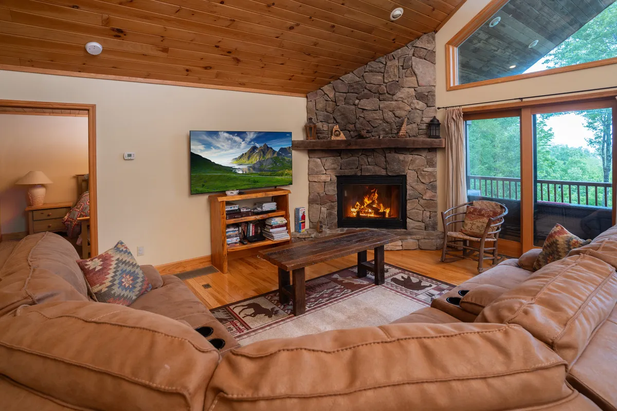 The spacious living room features a stunning stone fireplace at its heart, with a comfortable sectional sofa arranged for conversation. Large windows frame sweeping views of the surrounding mountains, flooding the space with natural light. The wooden coffee table and television add to the sense of a welcoming, multi-use gathering place.