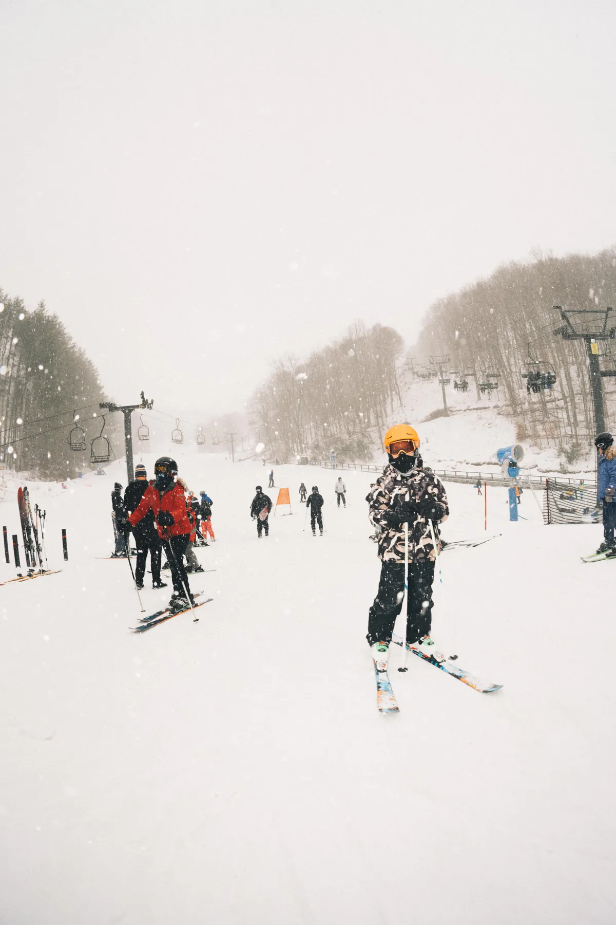A lively winter scene on the ski slopes, with skiers—including a child in a bright orange helmet—gliding down the snow, and the ski lift prominent in the background. The landscape is blanketed in crisp white snow, radiating energy, fun, and adventure.