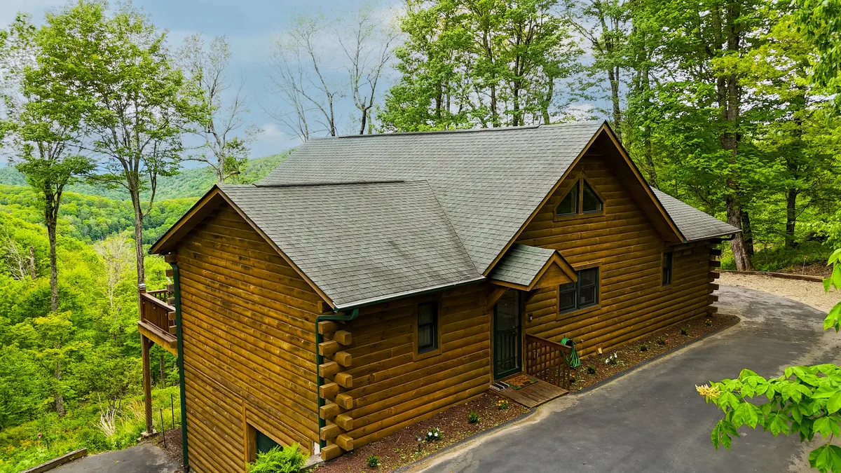 A classic log cabin is set among lush greenery, with a driveway leading up to its welcoming entrance. The cabin is tucked away, offering a sense of privacy and seclusion, while the surrounding landscape suggests peaceful walks and evenings under the stars.