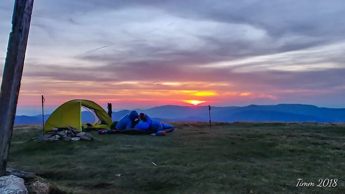 A couple relaxes together beside a tent, overlooking a spectacular mountain range as the sun sets behind the peaks. The scene is suffused with a romantic, golden glow, and the expansive view offers both intimacy and the drama of nature. The foreground shows the couple in a private moment, while the background highlights the immense beauty and seclusion of the mountains.