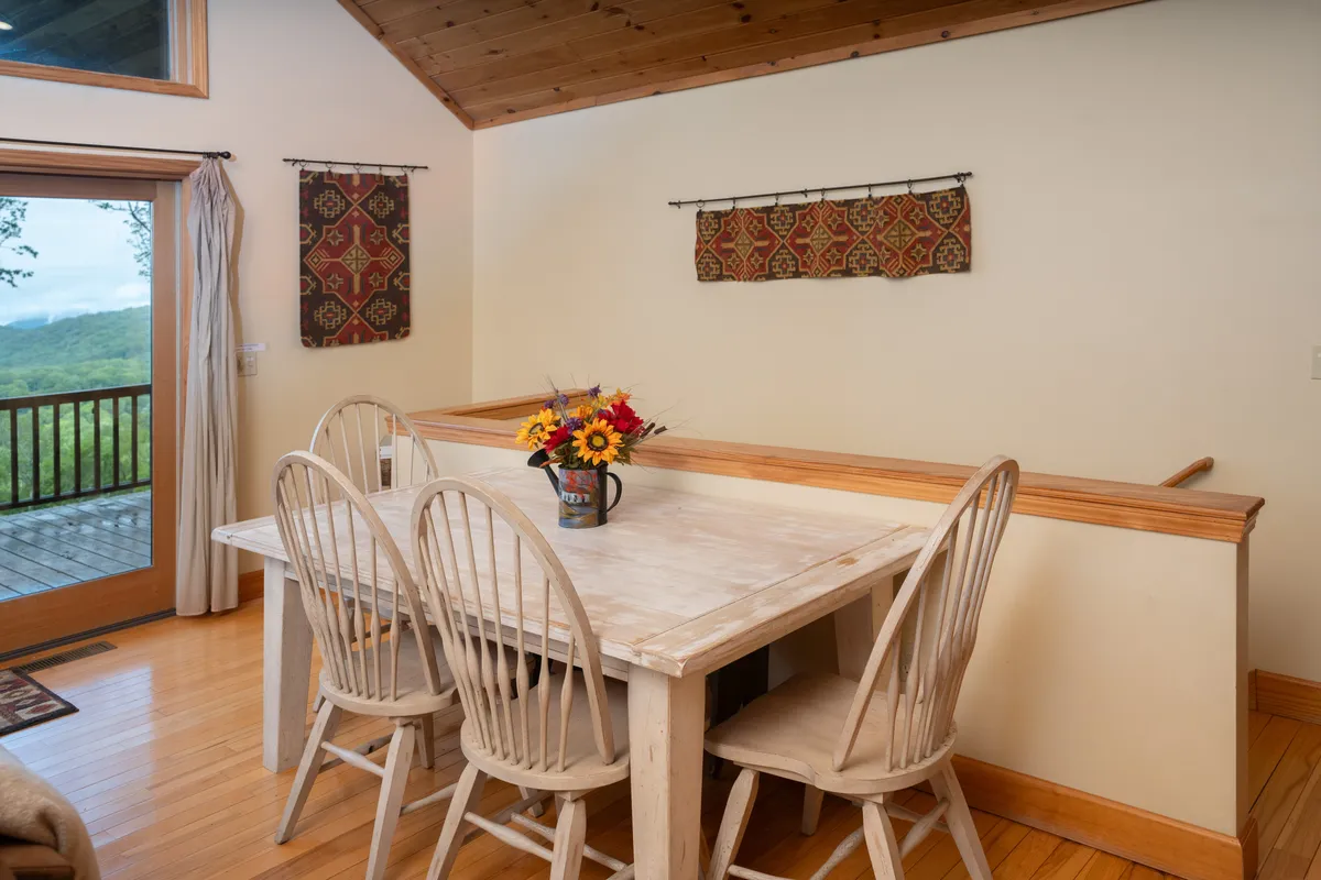 A cozy dining area with a wooden table set near a window, adorned with a flower vase. The glow from the outdoors illuminates the space, inviting guests to gather for meals or relaxed conversation while enjoying the scenery.