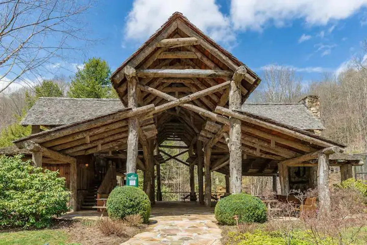 A charming log structure sits among landscaped gardens and a stone pathway, inviting groups to gather outdoors. The scene hints at spaces for relaxing, storytelling, or enjoying the garden ambiance, with a focus on the welcoming, communal spirit.
