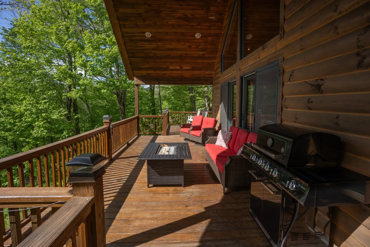 This image showcases a welcoming outdoor deck space with red cushioned chairs, a glass-top table, and a grill. The scene is set for relaxation or gathering, with ample seating and an open view, suggesting an ideal spot for unwinding and socializing. The atmosphere is inviting and relaxed, likely with scenic views beyond the deck railing.