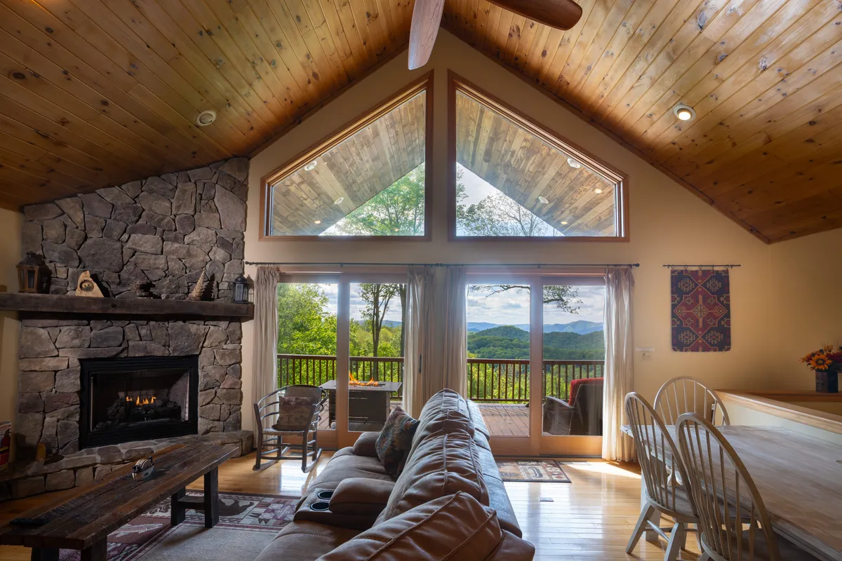 The interior image features a cozy living room with a stunning stone fireplace as the centerpiece, flanked by expansive windows that offer sweeping views of the mountains. A plush, comfortable sofa invites relaxation, while the warm wooden ceiling creates an inviting, rustic atmosphere perfect for unwinding. The natural light fills the space, highlighting the welcoming ambiance and the seamless connection to nature outside.